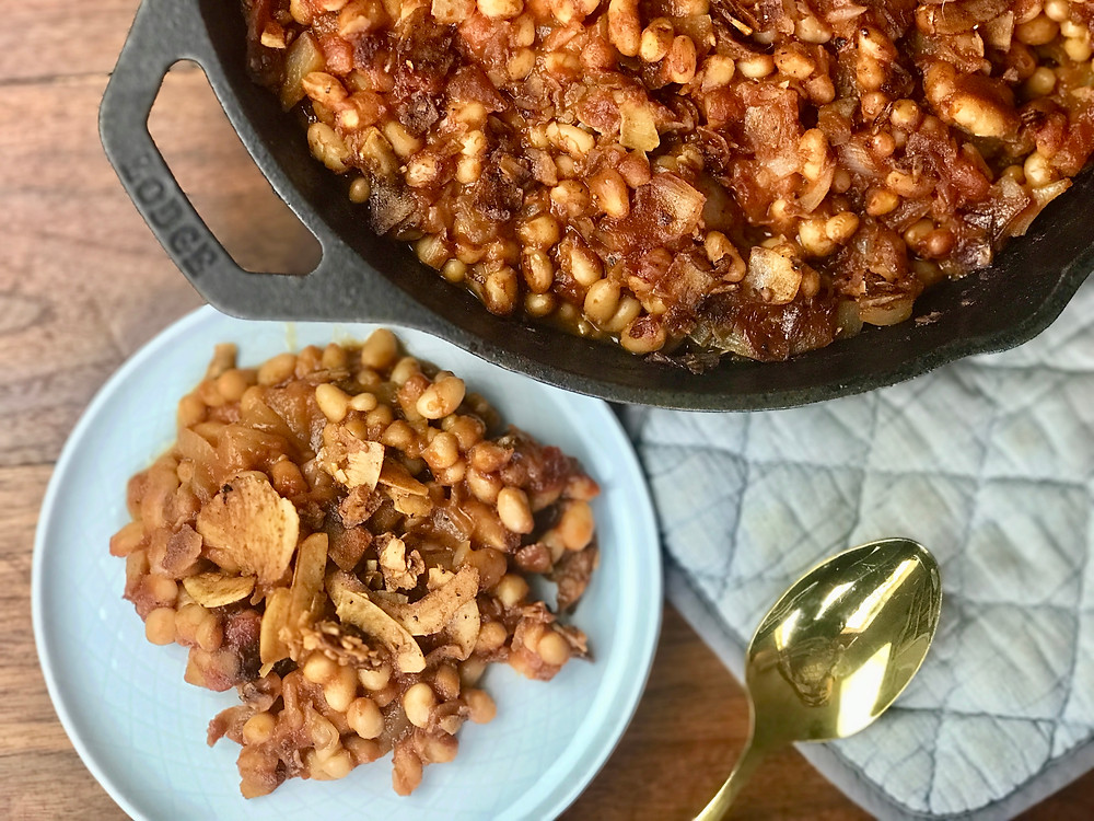 Cast iron skillet filled with saucy vegan baked beans topped with golden crispy coconut bacon